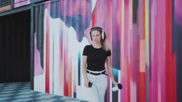 Милая молодая девушка танцует весело и беззаботно на фоне разноцветной стены. молодая женщина слушает музыку в наушниках на улице — стоковое видео