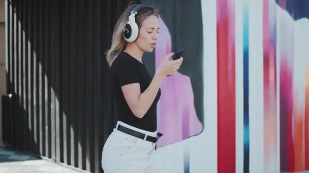 Portret van een aantrekkelijk meisje dat zorgeloos buiten danst. jonge vrouw in koptelefoon en met een papieren beker met koffie. — Stockvideo