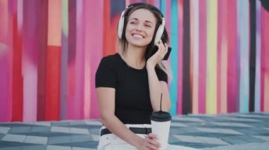Genç bir kadın çok renkli bir duvarın arkasında kaldırımda otururken kahve içiyor ve müzik dinliyor.
