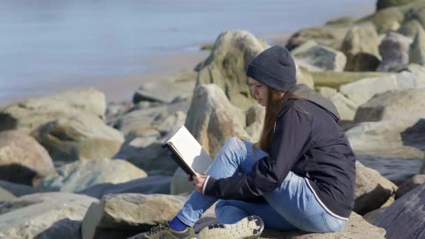 Jovem menina senta-se em uma praia rochosa e gosta do silêncio enquanto lê um romance — Vídeo de Stock