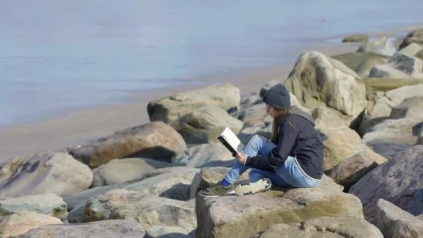 Chica joven se sienta en la playa con un libro en sus manos — Vídeo de stock