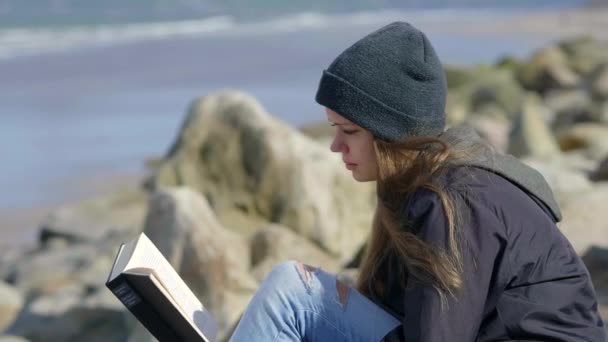 Mujer joven se sienta en el océano mientras lee un libro — Vídeo de stock