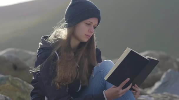 Mujer joven lee un libro mientras está sentada en las rocas — Vídeo de stock
