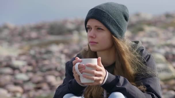 Menina bonita com uma xícara de café sentado em uma praia rochosa — Vídeo de Stock