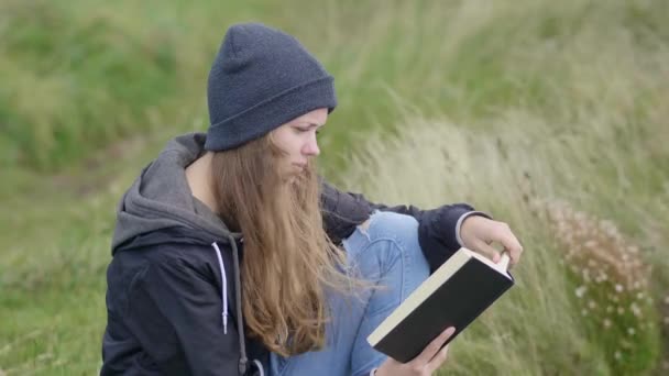 Menina senta-se na grama e lê um livro — Vídeo de Stock