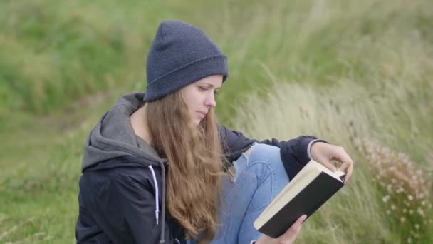 Jovem mulher senta-se na grama e lê um livro — Vídeo de Stock