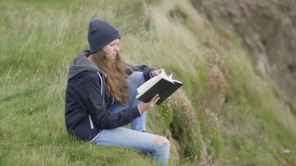 Chica se sienta en los campos de hierba en una colina mientras lee un libro — Vídeos de Stock