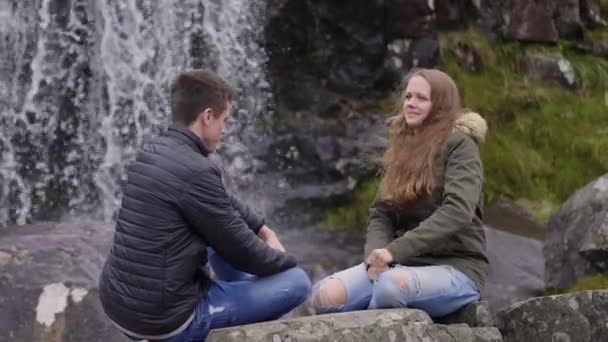 Dos amigos en un viaje de aventura a Irlanda sentados frente a una cascada — Vídeos de Stock