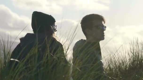 Ευτυχισμένο ζευγάρι κάθεται στο γρασίδι reed και απολαμβάνει το ηλιοβασίλεμα πάνω από τον ωκεανό — Αρχείο Βίντεο