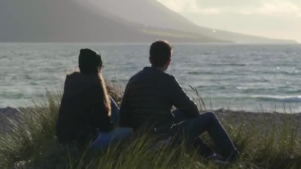 Pareja se sienta a la orilla del mar mientras observa la puesta de sol — Vídeo de stock