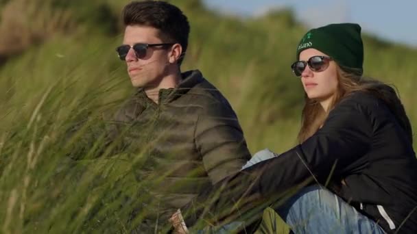 Молодая пара наслаждается закатом над океаном, сидя в тростниковой траве — стоковое видео