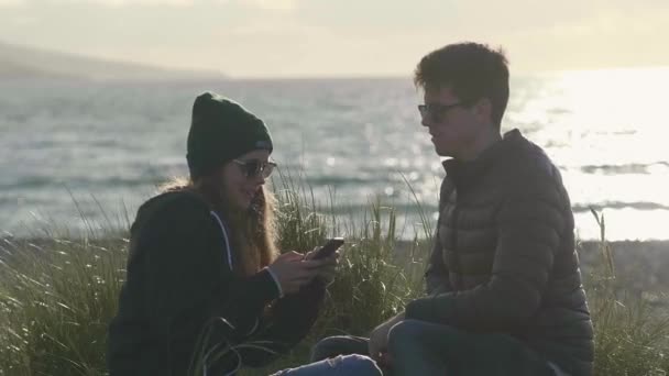 Bliska strzał z dwoma przyjaciółmi siedząc na plaży o zachodzie słońca — Wideo stockowe