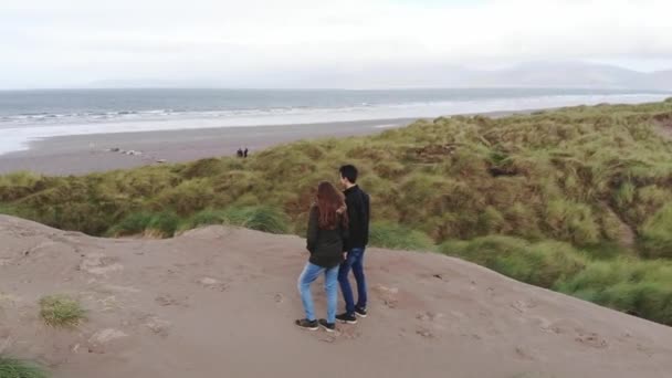 Voo em torno de um jovem casal em pé nas dunas de areia na costa — Vídeo de Stock