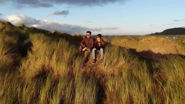 Vuelo de una joven pareja sentada en las colinas cubiertas de hierba en la costa — Vídeos de Stock