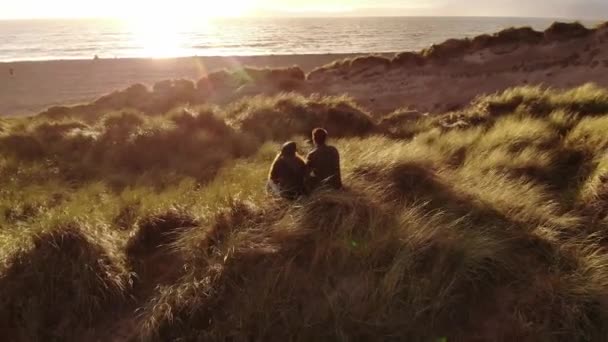 Flug um ein junges Paar, das bei Sonnenuntergang am Strand sitzt — Stockvideo