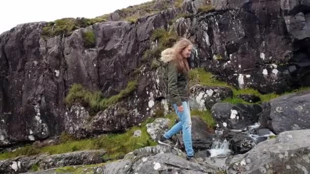 Молодая женщина перелезает через скалы перед водопадом — стоковое видео