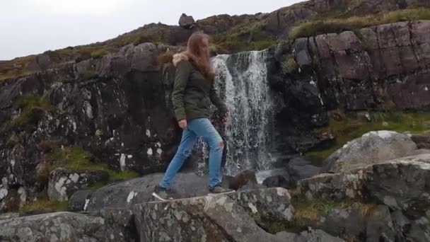 Une jeune femme grimpe sur des rochers devant une cascade — Video