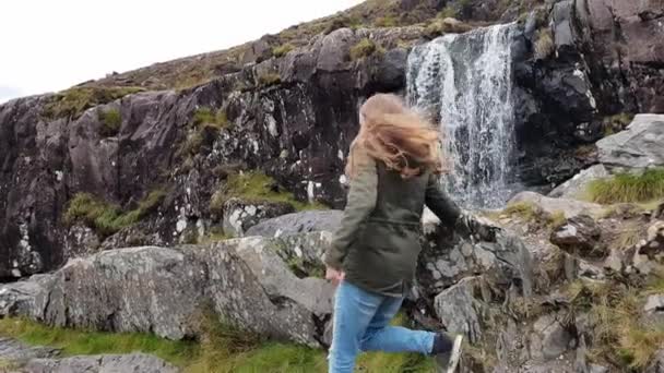 Młoda kobieta wspina się po skałach przy wodospadzie w Irlandii — Wideo stockowe
