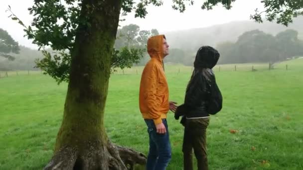 Pareja joven buscando refugio de fuertes lluvias bajo un árbol — Vídeos de Stock