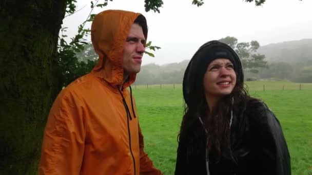 Молодая влюбленная пара ищет укрытия от дождя под деревом — стоковое видео