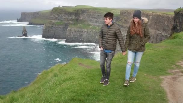 Jong koppel wandelingen hand in hand aan de rand van de beroemde Cliffs of Moher — Stockvideo