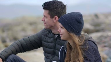 Genç çift okyanus üzerinde kayalık bir plajda otururken görünüyor