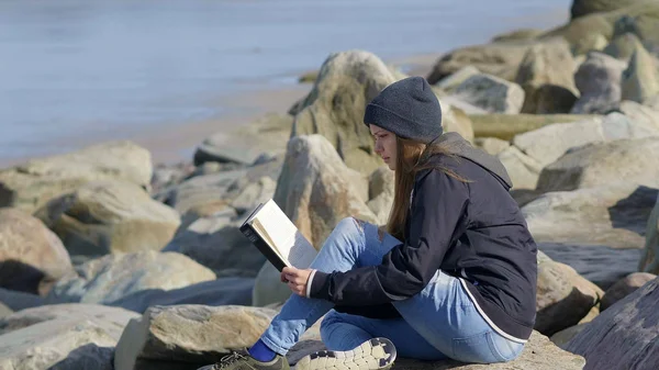 Giovane ragazza si siede su una spiaggia rocciosa e gode il silenzio durante la lettura di un romanzo — Foto Stock