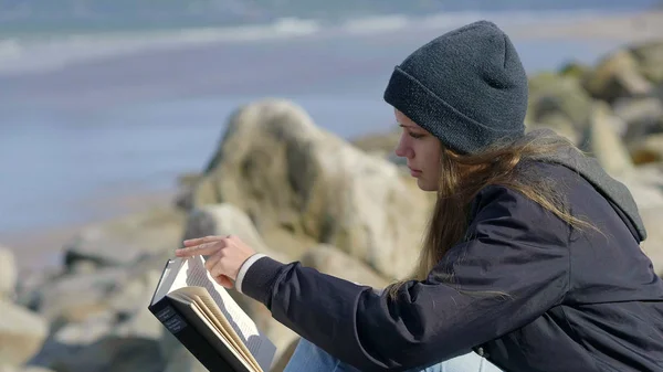 Young woman sits at the ocean while reading a book