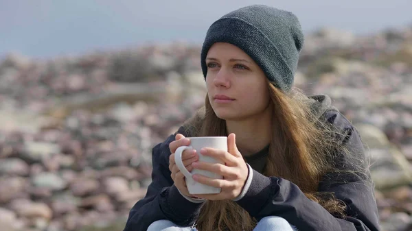 Menina bonita com uma xícara de café sentado em uma praia rochosa — Fotografia de Stock