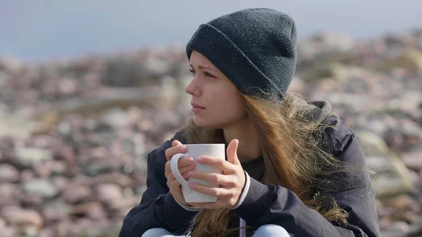 Krásná dívka s šálkem kávy sedí na skalnaté pláži — Stock fotografie