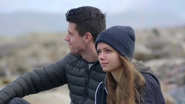 Young couple looks over the ocean while sitting on a rocky beach — Stock Photo, Image