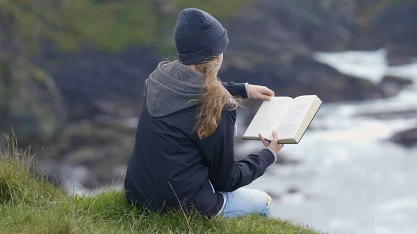 Rapariga senta-se à beira de um penhasco enquanto lê um livro — Fotografia de Stock