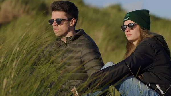 Young couple enjoys the sunset over the ocean while sitting in reed grass — Stock Photo, Image