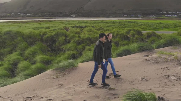 Pareja joven pasea por las dunas de arena en la costa irlandesa disfrutando de la vista sobre el océano — Foto de Stock