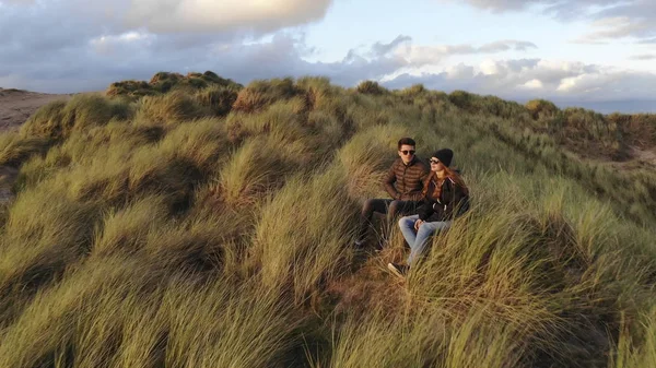 Vuelo hacia una joven pareja sentada en las colinas cubiertas de hierba en la costa — Foto de Stock