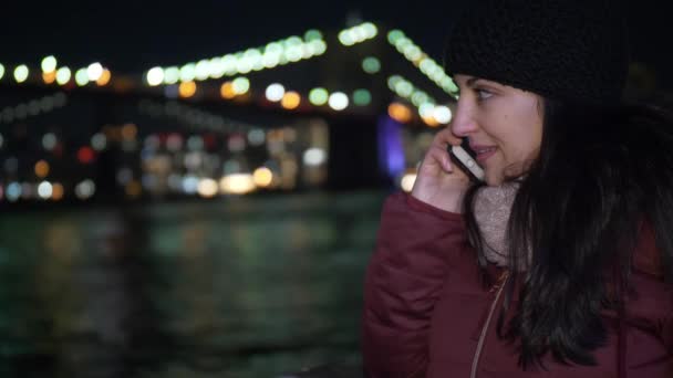 Jonge vrouw neemt een telefoontje op Brooklyn Bridge bij nacht — Stockvideo