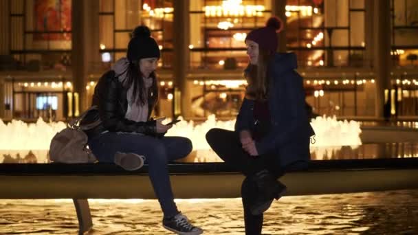 Två tjejer har en fantastisk kväll i New York när du sitter vid en fontän — Stockvideo