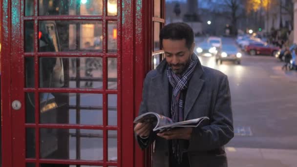 Homem na cidade de Londres lê uma revista enquanto se inclina contra uma cabine telefônica — Vídeo de Stock