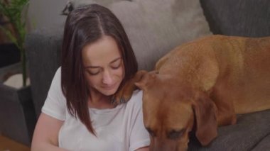 Genç bir kadın oturma odasında dinlenirken köpeğini okşar.