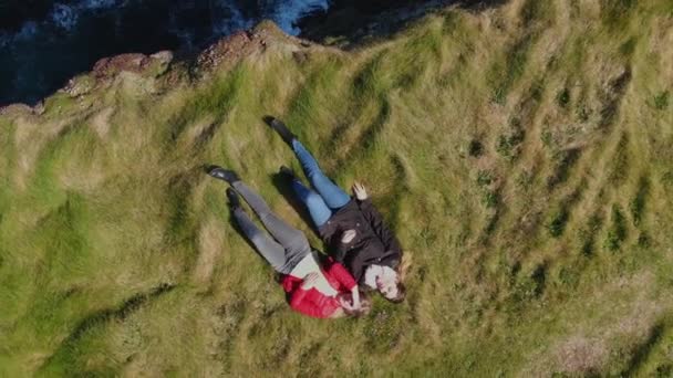 アイルランドの海岸の崖の草の中に横たわっている2人の女の子 上からドローンビュー — ストック動画