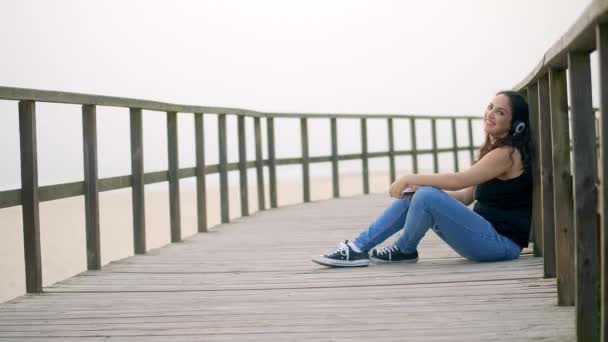 Jovem mulher senta-se em um cais de madeira na praia — Vídeo de Stock