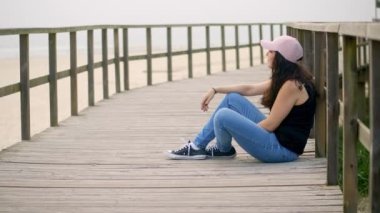 Genç bir kadın sahildeki ahşap iskelede oturuyor. Ağır çekimde.