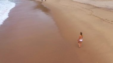 Bikinili seksi bir kadın okyanusta kumlu bir sahilde yürüyor. Hava aracı görüntüleri.