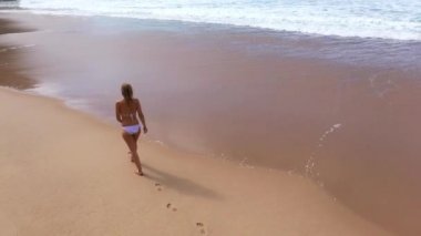 Bikinili seksi bir kadın okyanusta kumlu bir sahilde yürüyor. Hava aracı görüntüleri.