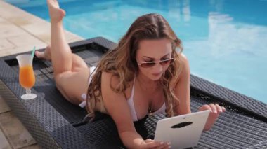 Bikinili genç bir kadın havuz kenarında tabletle rahatlıyor.