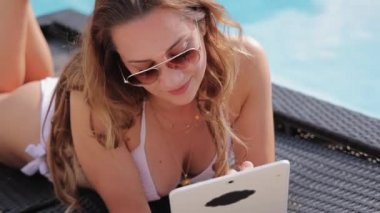 Bikinili genç bir kadın havuz kenarında tabletle rahatlıyor.