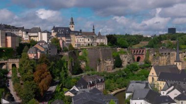 Lüksemburg şehrinin silueti bulutlu bir gökyüzü altında tarihi tahkimatlar, binalar ve yemyeşil alanları gösteriyor