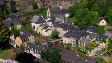 Lüksemburg 'un tarihi Grund bölgesinin panoramik manzarasında Alzette Nehri, renkli binalar, köprü ve yemyeşil alanlar sergileniyor.