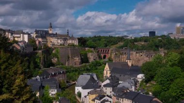 Lüksemburg 'un Grund bölgesinin panoramik manzarası, ortaçağ mimarisini, tahkimatlarını ve Neumunster Manastırı' nı dinamik bir gökyüzü altında sergiliyor.
