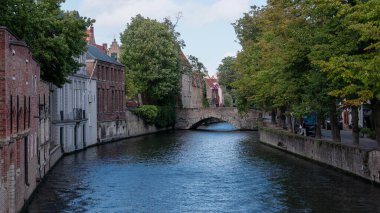 BRUGES, BELGIUM - 15 EYLÜL 2025 Bruges Kanalı eski binaların ve ağaçlarla çevrili taş bir köprünün önünden geçiyor